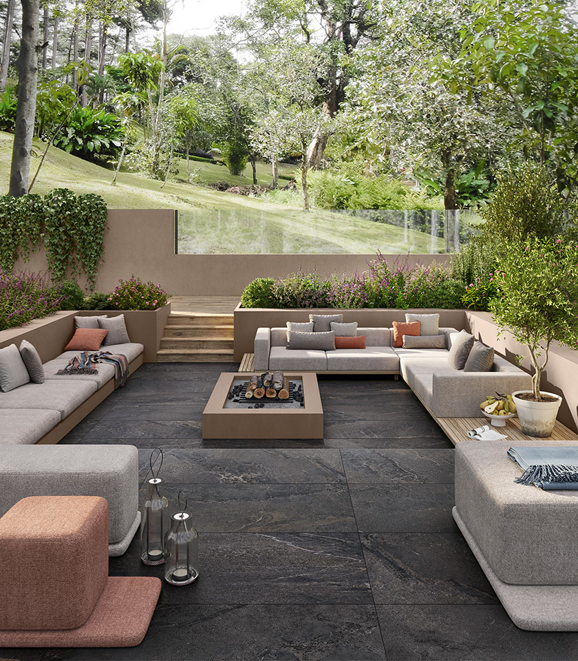 Outdoor living area surrounded by greenery with flooring from the Anthology collection in Dark Rough stone effect and elements from the Pigmenti collection in the Pumice shade.
