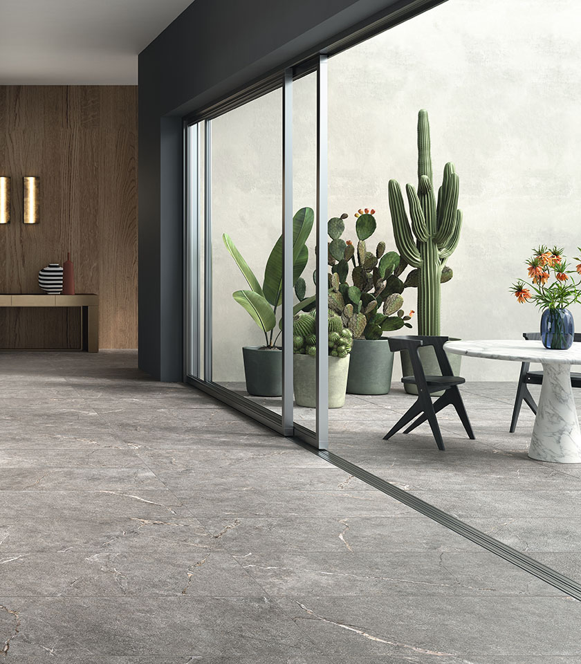 Contemporary space with grey stone-effect flooring extending seamlessly from indoors to outdoors, large glass doors, minimalist furniture and decorative plants.